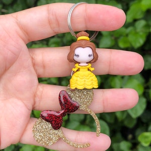 May include: Gold glitter Minnie Mouse ears with a red glitter bow and a Belle charm attached to a silver key ring.