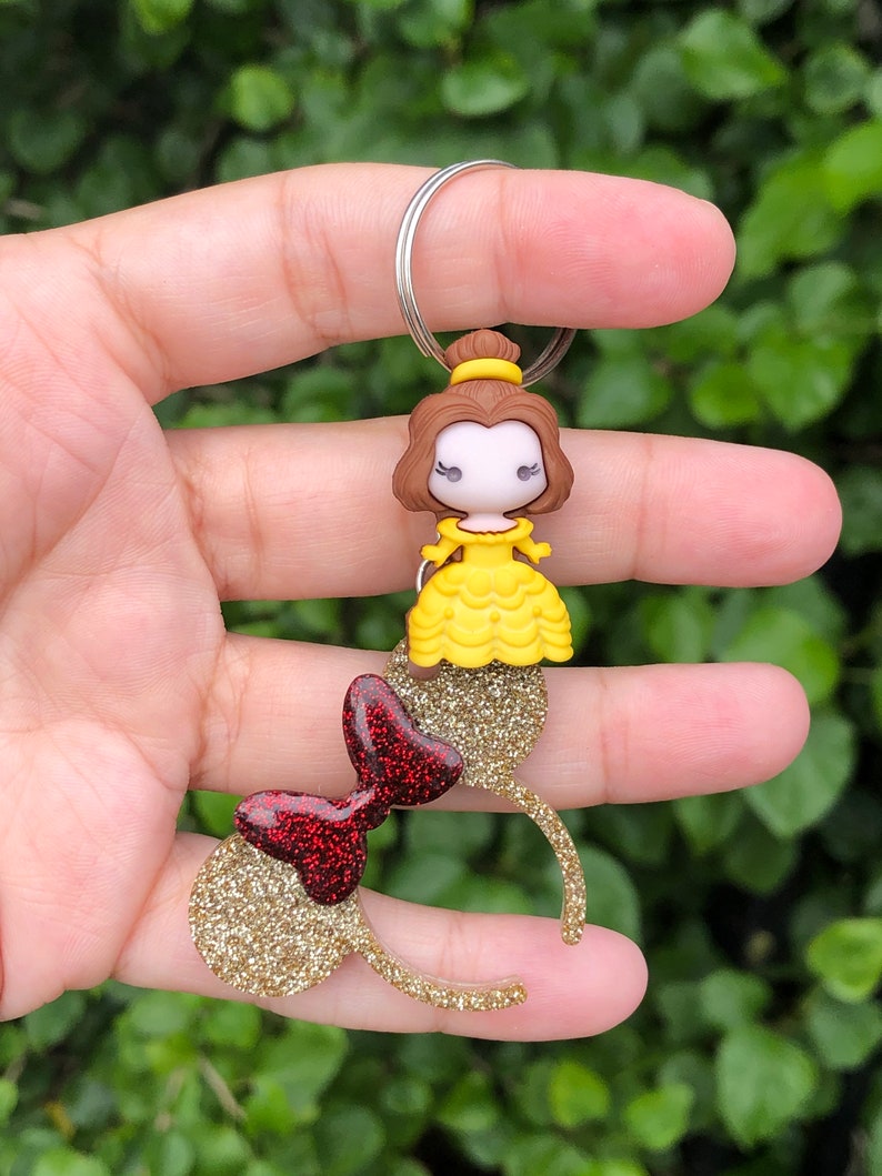 May include: Gold glitter Minnie Mouse ears with a red glitter bow and a Belle charm attached to a silver key ring.