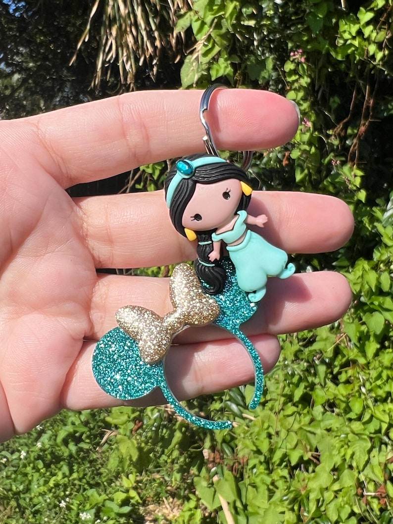May include: Teal blue and gold glitter Minnie Mouse ears with a small, teal blue and gold glitter Jasmine figurine attached.