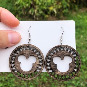 May include: Wooden dangle earrings featuring a dark brown circular design with a Mickey Mouse silhouette in the center. The earrings have a decorative openwork pattern around the mouse head and silver-colored ear wires.
