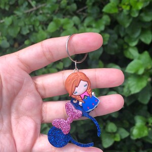 May include: Blue and pink glitter Minnie Mouse ears with a small, red-haired, blue-dressed character charm attached to the headband.