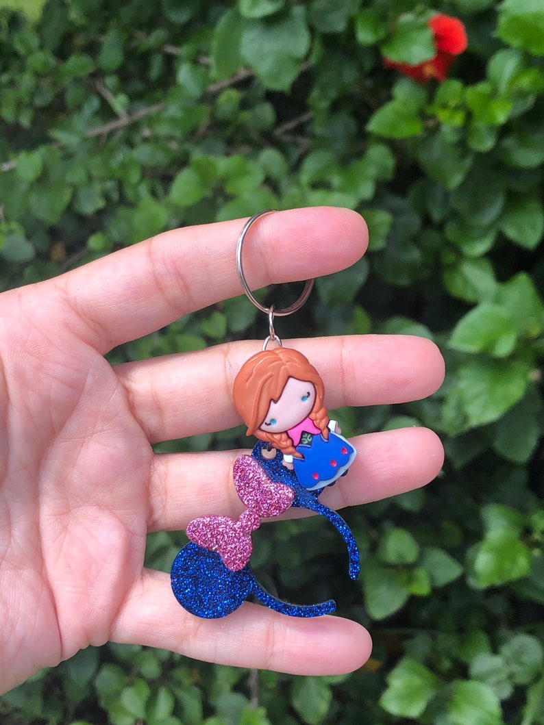 May include: Blue and pink glitter Minnie Mouse ears with a small, red-haired, blue-dressed character charm attached to the headband.