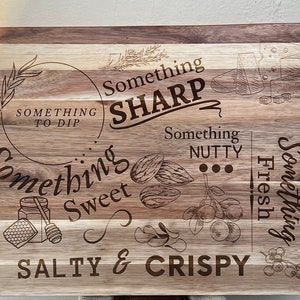 May include: A wooden cutting board with a light brown and dark brown wood grain pattern. The board is engraved with the text "Something Sharp", "Something Nutty", "Something Sweet", "Something Fresh", "Something to Dip", and "Salty & Crispy".