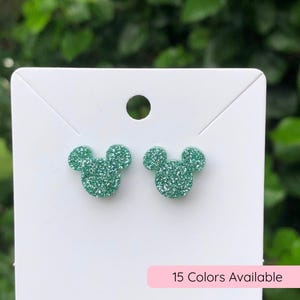 May include: A pair of mint green glitter stud earrings shaped like mouse heads attached to a white display card. The earrings feature a textured, sparkling surface. A small pink rectangular label at the bottom reads 15 Colors Available.