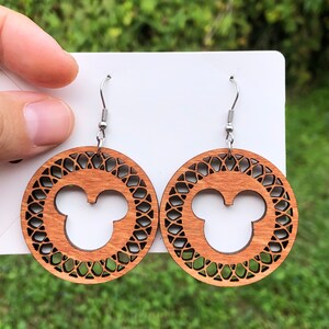 May include: Wooden earrings featuring a Mickey Mouse silhouette within a decorative circular frame. The earrings are a warm brown color and hang from silver-colored fish hook ear wires. The background is a blurred green.