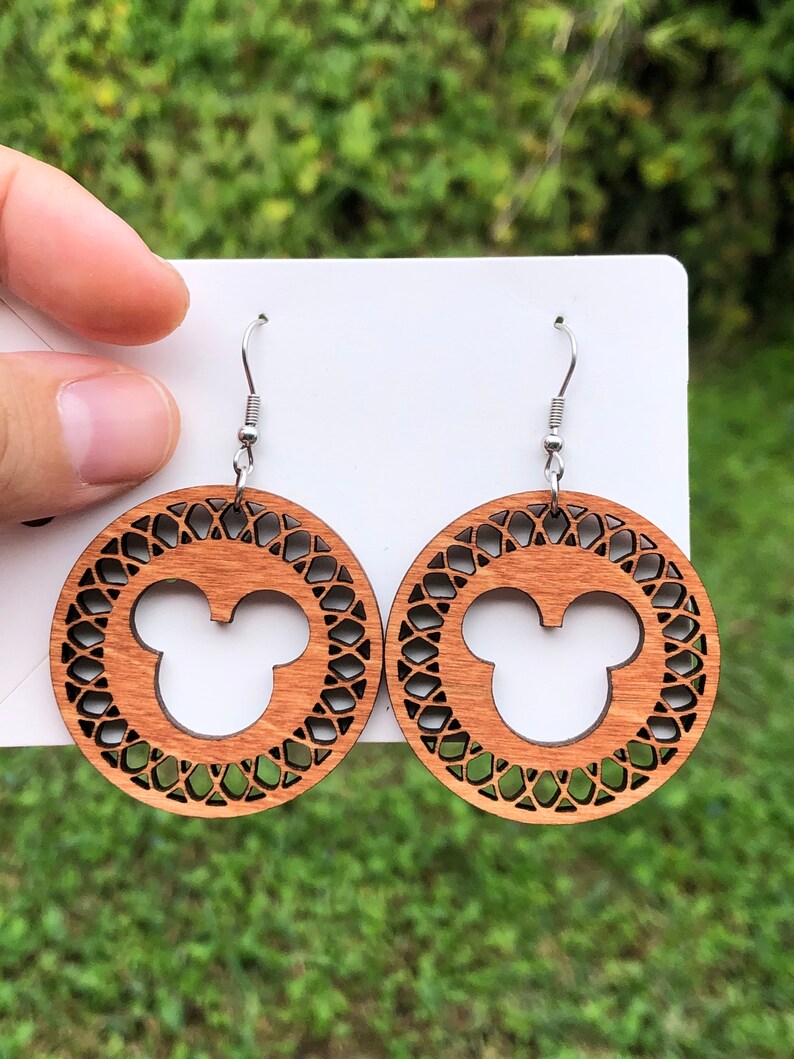 May include: Wooden earrings featuring a Mickey Mouse silhouette within a decorative circular frame. The earrings are a warm brown color and hang from silver-colored fish hook ear wires. The background is a blurred green.