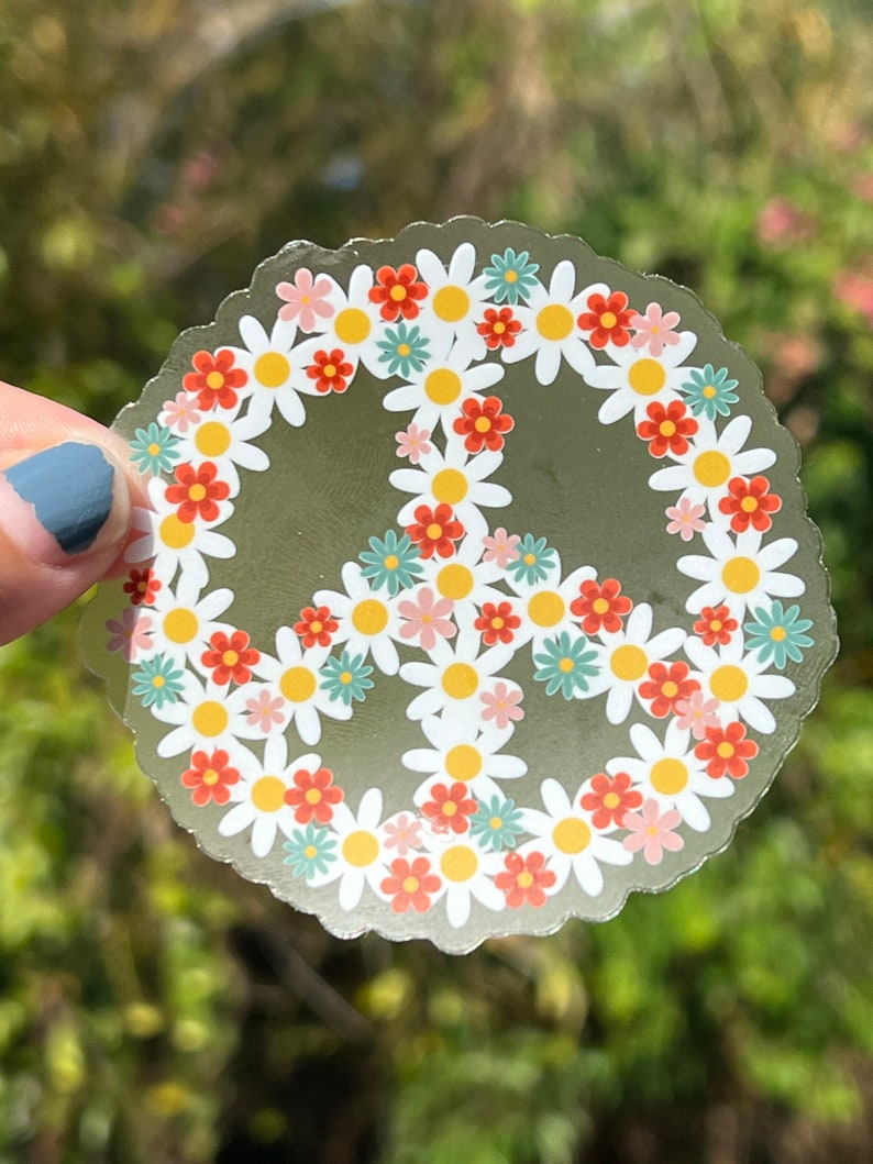May include: A clear, round sticker featuring a peace sign design. The peace sign is formed by a ring of white daisies with yellow centers, and interspersed with orange, pink, and teal flowers. The sticker has a scalloped edge.
