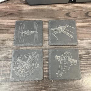 May include: Four square slate coasters with etched white designs. The designs include a TIE fighter, an X-wing, the Millennium Falcon, and Slave I. The coasters are arranged on a wooden surface.