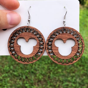 May include: A pair of wooden earrings featuring a circular design with a Mickey Mouse silhouette in the center. The earrings have a dark brown finish and intricate cut-out patterns around the edges, with silver-colored ear wires.