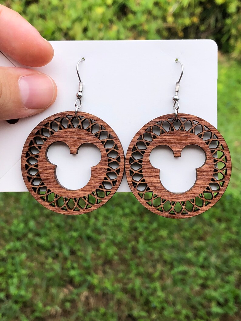 May include: A pair of wooden earrings featuring a circular design with a Mickey Mouse silhouette in the center. The earrings have a dark brown finish and intricate cut-out patterns around the edges, with silver-colored ear wires.