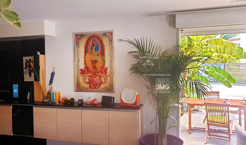 Puede incluir: Una impresi&oacute;n enmarcada de una imagen religiosa colorida con una mujer con un vestido azul y un fondo rojo. La impresi&oacute;n est&aacute; colgada en una pared blanca en una habitaci&oacute;n con una planta grande y una ventana que da a un patio.