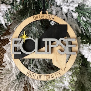 May include: A wooden ornament with a black and gray silhouette of the path of totality for the 2024 solar eclipse. The ornament is inscribed with "ECLIPSE" and "PATH OF TOTALITY" and the date "04.08.2024".