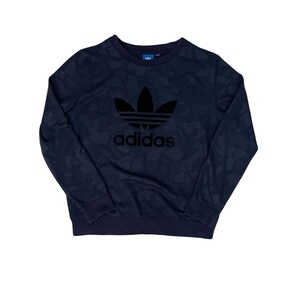 Könnte beinhalten: Marineblaues Sweatshirt mit Camouflage-Muster. Das schwarze Adidas-Logo ist prominent auf der Vorderseite zu sehen. Das Sweatshirt hat einen Rundhalsausschnitt und lange Ärmel. Das Adidas-Logo ist ein Trefoil.