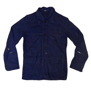 Peut inclure: Veste de travail bleu marine vintage avec une fermeture boutonnée, un col et plusieurs poches. La veste présente des signes d'usure, avec des traces de peinture et une légère décoloration. Les manches longues s'étendent jusqu'aux poignets.