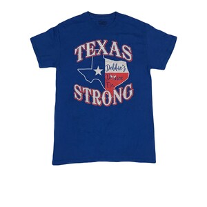 Op de afbeelding: Een koninklijk blauw T-shirt met een witte omtrek van de staat Texas en de woorden "Texas Strong" in wit met rode contouren. Binnen de omtrek van Texas bevindt zich een rood-wit-blauwe Amerikaanse vlag met de woorden "Debbie's Dance Etc." in wit.