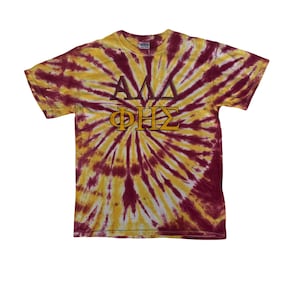 May include: A tie-dye t-shirt in shades of yellow and maroon. The shirt features the Greek letters ΑΛΔ above ΦΗΣ in a contrasting brown color. The t-shirt is a short-sleeved crew neck.