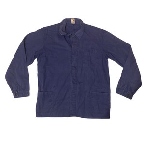 Può includere: Giacca da lavoro vintage blu navy con maniche lunghe, due tasche anteriori e una piccola tasca sul petto. La giacca ha una chiusura con bottoni e un colletto. Il tessuto mostra segni di usura. Una piccola etichetta è visibile sul colletto.