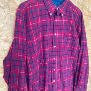 May include: A red, blue and white plaid long-sleeved button-up shirt. The shirt has a pocket on the left side and a label that reads "Made in U.S.A."