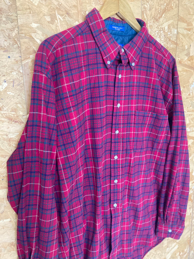 May include: A red, blue and white plaid long-sleeved button-up shirt. The shirt has a pocket on the left side and a label that reads "Made in U.S.A."