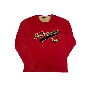 May include: A red long-sleeved top with the text "San Francisco" in yellow script and a navy blue underline. The top also features the letters "LSC" in gold. The top has a crew neck and a light tan lining.