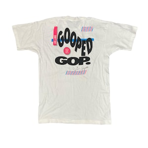 Puede incluir: Camiseta blanca con un gráfico negro y azul que dice "Goop'd GOP?" con un círculo rojo que dice "Up Off It" y un patrón de rayas azules y rojas.