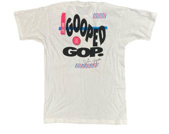 May include: White t-shirt with a black and blue graphic that says "Goop'd GOP?" with a red circle that says "Up Off It" and a blue and red striped pattern.