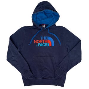 May include: Navy blue hooded sweatshirt with a blue interior lining and a front pocket. The front features the text "THE NORTH FACE" in red, blue, and white. The hood has a blue drawstring.