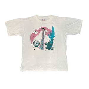 Peut inclure: T-shirt blanc avec un motif de désert du Sud-Ouest. Le graphisme comprend une femme en robe longue, un cactus turquoise et un paysage rose et blanc. Le motif comprend également un pot décoratif et un motif géométrique.