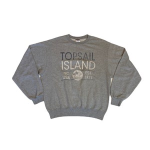 Può includere: Una felpa grigio mélange con la scritta "TOPSAIL ISLAND" in blu navy e grigio chiaro. Sotto, "NC USA EST. 1971" con un disegno di una palma e del sole. La felpa ha maniche lunghe e girocollo.