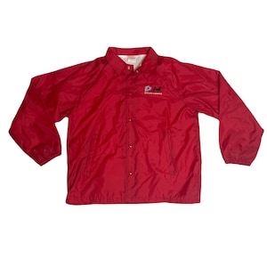 Puede incluir: Una chaqueta de nylon roja con cierre de presión y mangas largas. La chaqueta tiene un logotipo bordado en el pecho izquierdo que dice "Dover Downs".