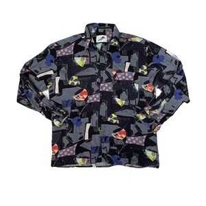 May include: A long-sleeved shirt with a distinctive abstract design. The shirt has a dark base colour with grey, blue, yellow, pink, and white geometric shapes. The collar and cuffs are visible, and the shirt is unbuttoned.