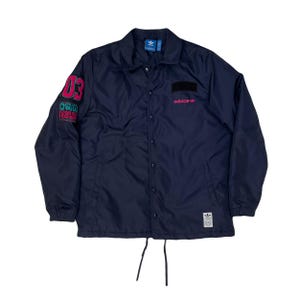 May include: Navy blue Adidas coach jacket with a button-up front and a collar. The jacket features the Adidas logo in pink on the chest and a graphic on the left sleeve with the number "03". A small Adidas logo patch is on the bottom.