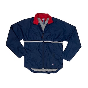 May include: A navy blue zip-up jacket with a red collar and white stripe across the chest. The jacket has a small logo on the chest and is made of a lightweight material. The jacket is a vintage style.