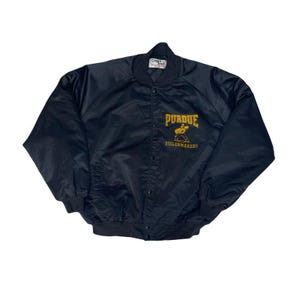 May include: A black satin bomber jacket with a snap-front closure. The jacket features the Purdue Boilermakers logo in gold on the chest. The jacket has a ribbed collar, cuffs, and waistband.