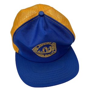 May include: A blue and gold trucker hat with a curved brim. The hat features a blue front panel with a gold emblem and text. The mesh back is gold. The hat is a vintage style.