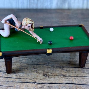 May include: A miniature green felt pool table with a brown wooden frame and gold accents. A figurine of a woman in a black dress is taking a shot with a pool cue. There are three balls on the table: a red ball, a white ball, and a black ball.