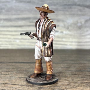 Op de afbeelding: Miniatuurbeeldje van een man in een sombrero en poncho, met een pistool en een klein groen voorwerp. Het figuur draagt witte en bruine kleding, met bruine laarzen. Het beeldje staat op een houten voet.