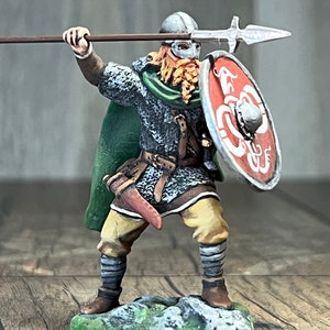 Könnte beinhalten: Eine bemalte Miniaturfigur eines Wikingerkriegers, der eine Lanze und einen Schild hält. Der Krieger trägt einen grünen Umhang, eine Kettenrüstung und einen Helm mit einer rot-weißen Gesichtsmaske. Der Schild ist rot mit einem weißen Drachenmuster.