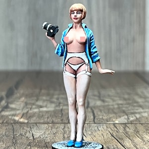 May include: A figurine of a woman wearing a blue shirt, white lingerie, and white stockings. She is holding a camera in her right hand and has blonde hair.