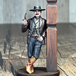 May include: A painted miniature figurine of a man wearing a black hat, a dark jacket, and brown boots. He is holding a pipe in his right hand and leaning against a wooden post.