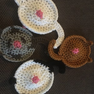 Puede incluir: Cuatro posavasos de ganchillo con forma de trasero de gato. Cada posavasos es de un color diferente: blanco, gris, marr&oacute;n y blanco con detalles negros. Cada posavasos tiene un c&iacute;rculo de ganchillo rosa en el centro.