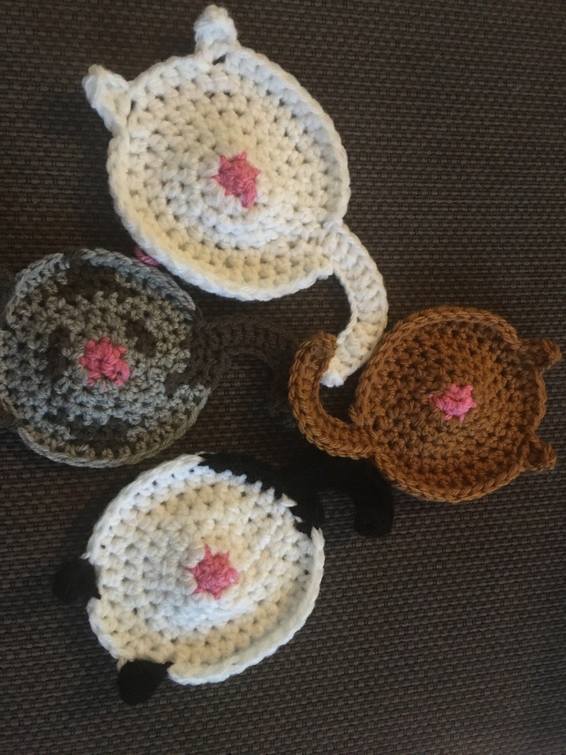 Puede incluir: Cuatro posavasos de ganchillo con forma de trasero de gato. Cada posavasos es de un color diferente: blanco, gris, marr&oacute;n y blanco con detalles negros. Cada posavasos tiene un c&iacute;rculo de ganchillo rosa en el centro.