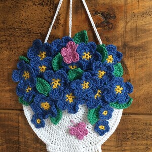 May include: A crocheted hanging planter with blue flowers, green leaves, and pink butterflies. The planter is white and has a round shape.