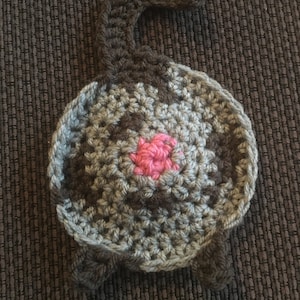 Puede incluir: Un juguete de gato de crochet con un cuerpo gris y marr&oacute;n y una nariz rosa. El gato est&aacute; acostado de espaldas con la cola enroscada.
