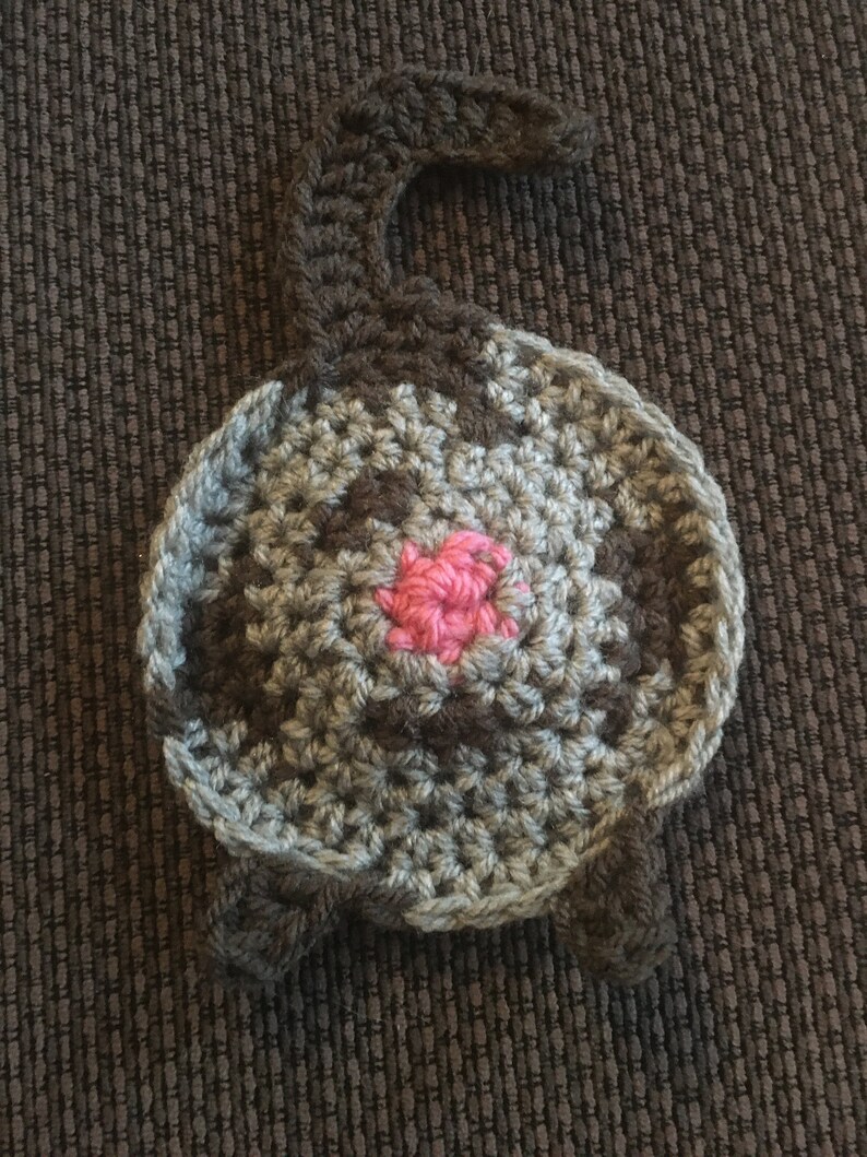Puede incluir: Un juguete de gato de crochet con un cuerpo gris y marr&oacute;n y una nariz rosa. El gato est&aacute; acostado de espaldas con la cola enroscada.
