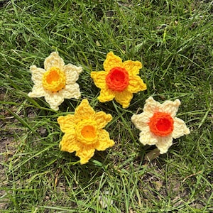 May include: Four handmade crochet daffodil flowers in shades of yellow, cream, and orange, arranged on green grass. The flowers have detailed petals and a textured appearance, showcasing the craftsmanship. A charming, spring-themed craft.