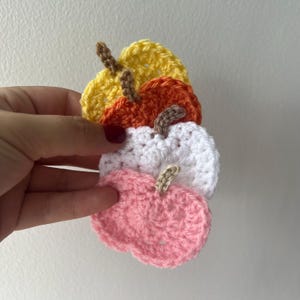 Könnte beinhalten: Vier gehäkelte Kürbisse in Gelb, Orange, Weiß und Rosa, jeder mit einem braunen Stiel. Diese handgefertigten Kürbisse sind wahrscheinlich für Herbstdekorationen oder Bastelprojekte gedacht. Die Kürbisse sind gestapelt.