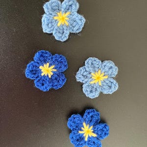 Peut inclure: Quatre fleurs faites à la main au crochet, dans des tons de bleu avec des centres jaunes. Les fleurs sont disposées sur une surface sombre. Les pétales sont détaillés et les centres jaunes ajoutent un contraste vif au bleu.
