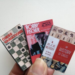 May include: Three miniature hardcover books about chess. The first book is titled "Chess Variations" and features a chessboard on the cover. The second book is titled "Bobby Fischer" and features a black and white photo of Bobby Fischer on the cover. The third book is titled "A History of Chess" and features a chessboard with chess pieces on the cover.