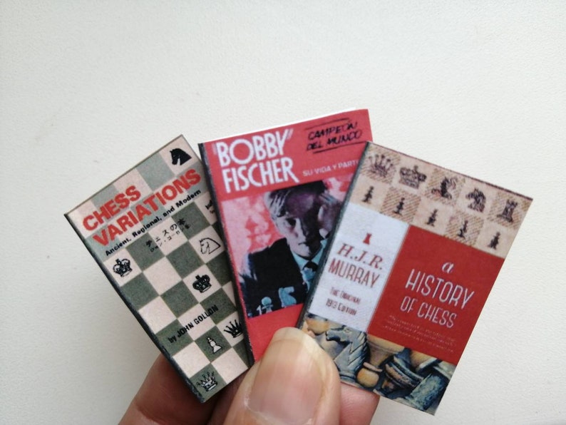 May include: Three miniature hardcover books about chess. The first book is titled "Chess Variations" and features a chessboard on the cover. The second book is titled "Bobby Fischer" and features a black and white photo of Bobby Fischer on the cover. The third book is titled "A History of Chess" and features a chessboard with chess pieces on the cover.